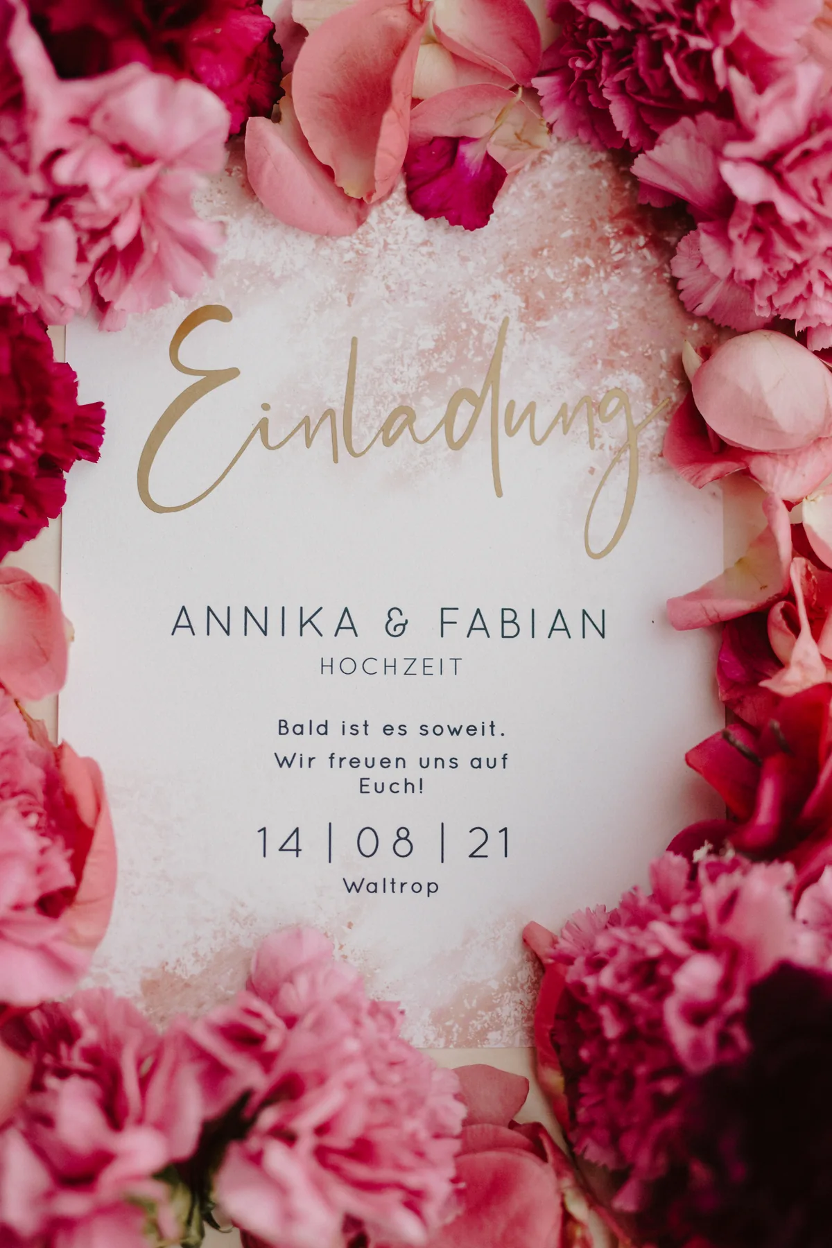 Hochzeitseinladung in Rosé und Pink mit goldener Kalligrafie, umgeben von Nelken